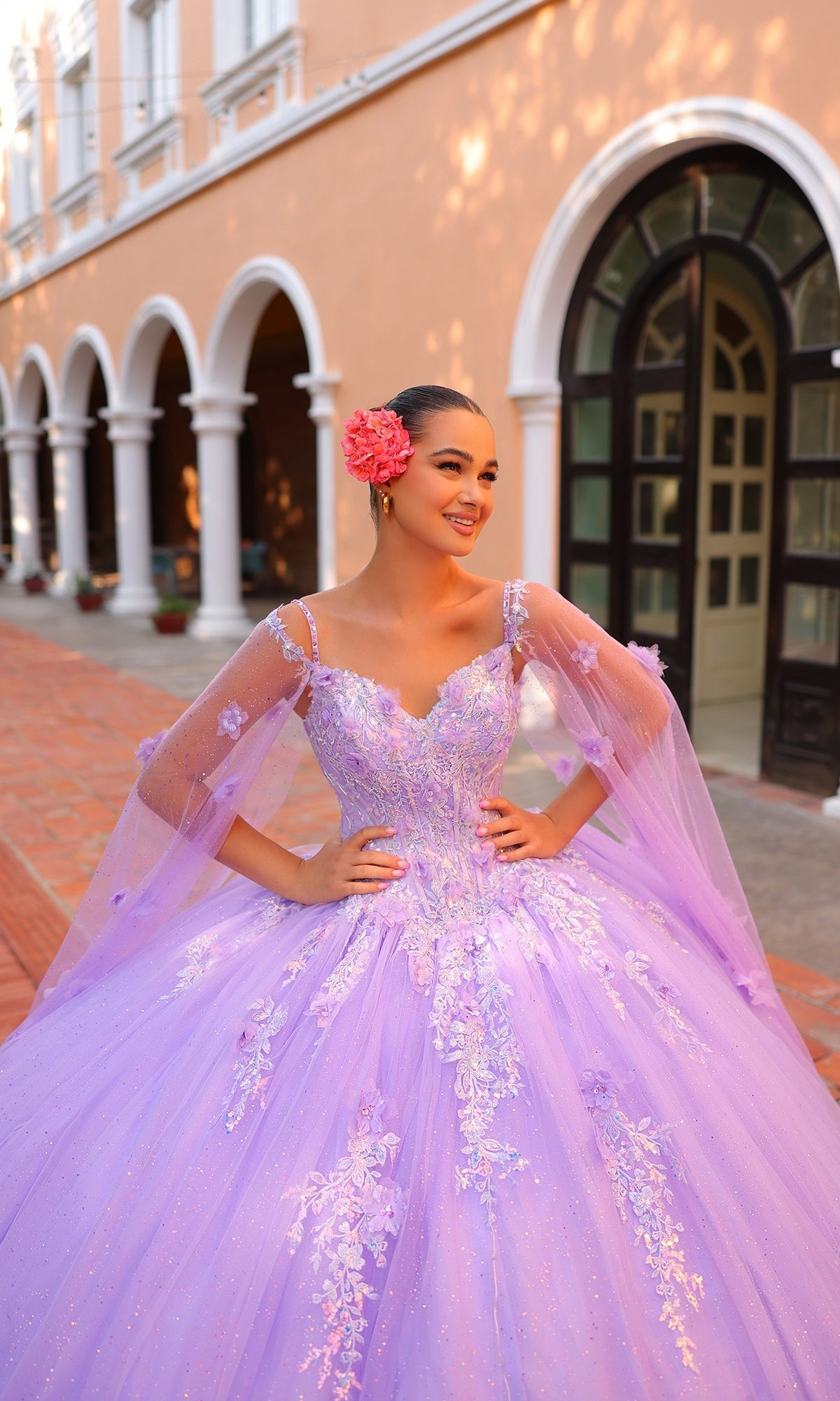 A young woman in the Quinceanera Dress 54306 By Amarra, featuring floral embroidery and a corset bodice in lavender, stands smiling outside an arched building with a pink flower in her hair.